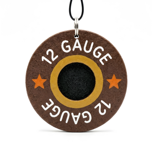 A round wooden charm designed to look like a 12 gauge shotgun shell, with a black primer center, gold ring, and orange stars on a brown background, hanging from a black cord.