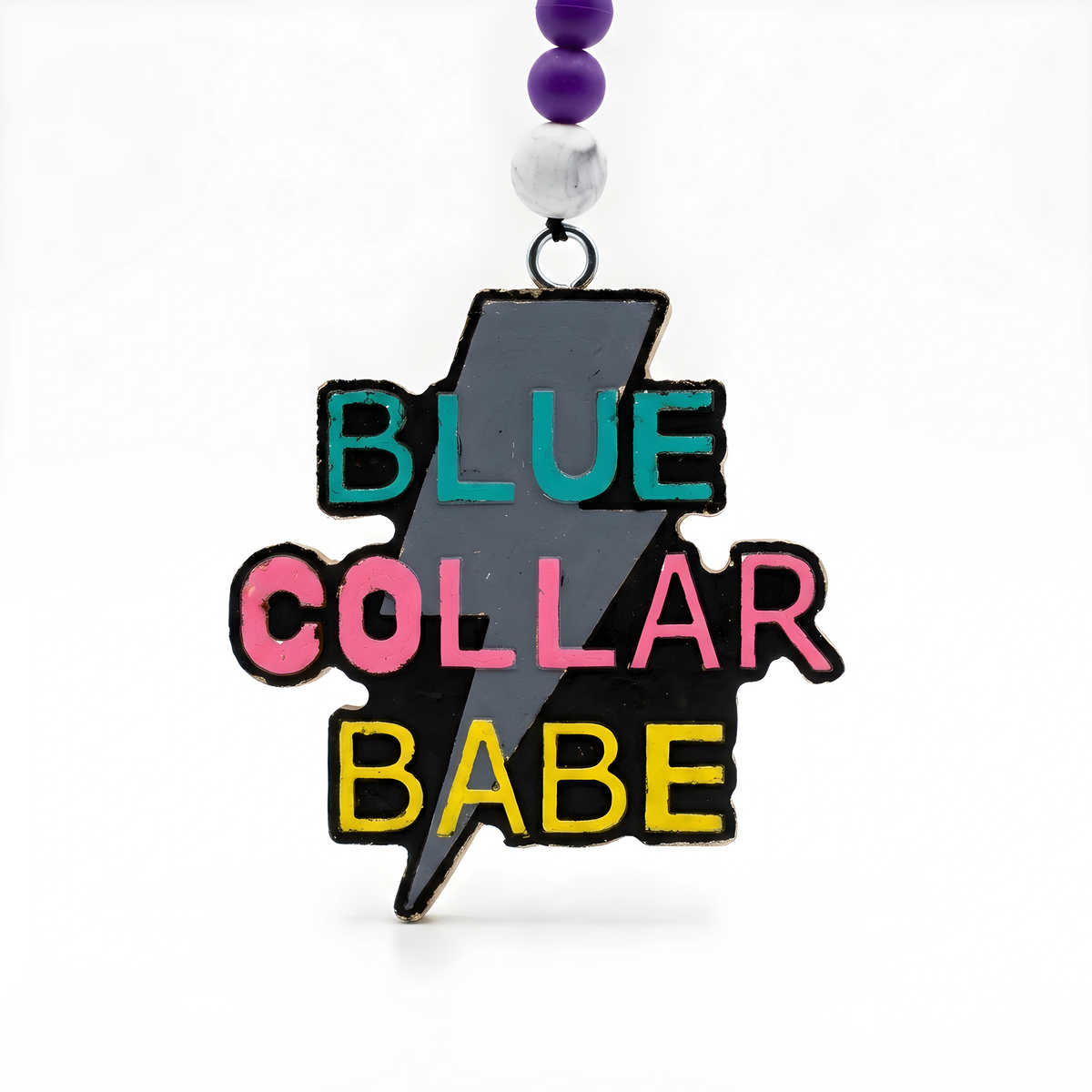 A lightning bolt-shaped charm with the words BLUE COLLAR BABE in teal, pink, and yellow lettering on a black background, hanging from a black cord with purple and white beads.