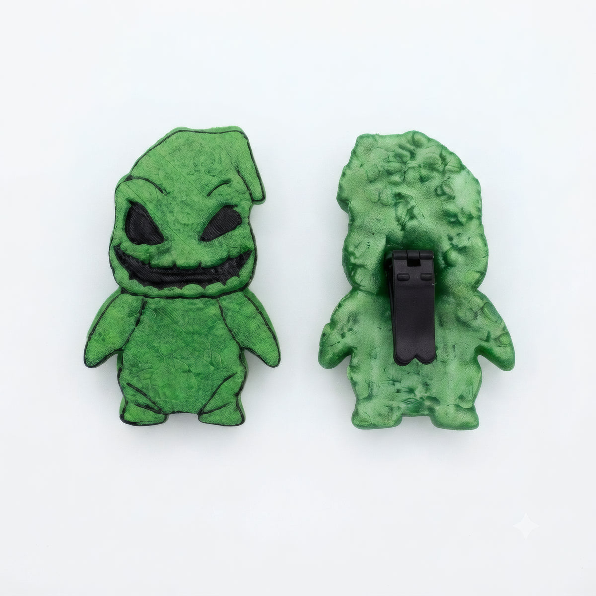 Two green character-shaped accessories shown on a plain white background, one front view and one back view with a black clip fastener visible.